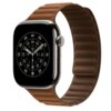 Na zdjęciu widoczny jest smartwatch z okrągłą tarczą wyświetlającą cyfry i oznaczenia minutowe, zamocowany na karmeowym pasku o teksturze z wyraźnymi przeszyciami. Pasek jest przymocowany do zegarka za pomocą magnetycznego zapięcia.