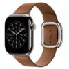 Karmelowy pasek do zegarka Apple Watch z klamrą, przymocowany do koperty zegarka z wyświetlaczem analogowym. Tarcza zegarka pokazuje cyfry i oznaczenia minutowe, a pasek ma kilka dziurek do regulacji.