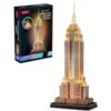 Na zdjęciu znajduje się opakowanie puzzli 3D oraz złożony model Empire State Building w wersji nocnej, oświetlony ciepłym światłem. Na opakowaniu widnieje napis: "CubicFun New York Empire State Building Night Edition 37 Pieces 3D Puzzle".