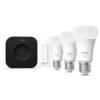 Na zdjęciu znajduje się zestaw startowy Philips Hue, składający się z trzech żarówek, mostka oraz regulatora. Na żarówkach widoczny jest napis "hue white and color 1100", a na mostku i regulatorze logo "hue".
