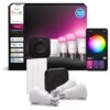 Na zdjęciu znajduje się zestaw startowy Philips Hue, który obejmuje trzy żarówki, mostek, regulator oraz opakowanie z logo Philips Hue i oznaczeniami kompatybilności z systemami Alexa, Apple Home, Google Home i Samsung SmartThings. Obok zestawu widoczny jest smartfon z aplikacją do sterowania oświetleniem, pokazującą koło barw i opcje regulacji.