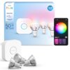 Na zdjęciu znajduje się zestaw trzech inteligentnych żarówek LED PHILIPS HUE 929004235707 4.7W GU10, opakowanie z napisem "PHILIPS hue Essential GU10 Starter kit White and color ambiance 50W 345 lumen" oraz aplikacja na smartfonie z kołem kolorów. Obok widoczny jest biały mostek z logo PHILIPS.