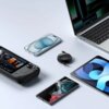 Na zdjęciu widoczne są różne urządzenia elektroniczne, w tym laptop, smartfony, tablet oraz kontroler do gier, a także kabel USB-C - USB-C BASEUS Free2Pull Mini 100W 1.5 m Czarny. Kabel jest zwinięty w okrągły kształt z widocznym logo "Baseus" na obudowie