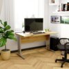 Na zdjęciu znajduje się biurko BM-135 Dąb Artisan o wymiarach 135x60x76 cm, na którym stoi monitor, klawiatura i myszka. W tle widać roślinę doniczkową, krzesło biurowe oraz półki z książkami i dekoracjami.
