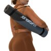Szaro-czarna mata do jogi z napisem "eneroFIT" jest zwinięta i przewieszona przez ramię osoby ubranej w brązowy strój sportowy. Mata posiada czarny pasek do noszenia.