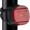 Czarno-czerwona lampka rowerowa zamocowana na pionowej rurce, z widocznym czerwonym kloszem i oznaczeniem "K 2234". Produkt ma kompaktowy, prostokątny kształt z zaokrąglonymi rogami.