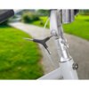 Na zdjęciu widoczny jest fragment roweru z zamontowanym kluczem rowerowym w kształcie litery T, przykręconym do śruby na kierownicy. W tle znajduje się rozmyty widok zielonego trawnika i ścieżki.