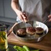Na zdjęciu widać osobę używającą metalowej szpatułki do przewracania kotleta mielonego na patelni, obok której stoi butelka z olejem oraz leżą zielone papryki. Patelnia znajduje się na drewnianej desce do krojenia.