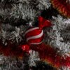Na zdjęciu widoczna jest czerwona bombka w kształcie cukierka z białymi, błyszczącymi paskami, zawieszona na ośnieżonej choince. W tle znajduje się czerwona i złota girlanda.