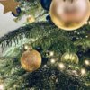 Na zdjęciu widoczna jest choinka ozdobiona złotymi i zielonymi bombkami oraz światełkami. W tle znajduje się złota gwiazda, a gałęzie choinki są gęsto pokryte igłami.