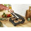 Na zdjęciu widoczny jest grill elektryczny z otwartą pokrywą, na którym znajdują się cztery kawałki mięsa z widocznymi śladami grillowania. W tle na blacie kuchennym znajdują się różne warzywa, przyprawy oraz deski do krojenia.