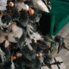 Na zdjęciu znajduje się ozdobiona choinka z ciemnozielonymi gałązkami, na której wiszą złote i miedziane bombki oraz białe dekoracje. Obok choinki stoi zielona sofa, a na podłodze leżą zapakowane prezenty z kokardami.
