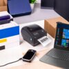 Na zdjęciu znajduje się drukarka z etykietą "Zenwire", z której wychodzi wydruk z kodem kreskowym, obok leży smartfon podłączony kablem oraz laptop z widocznym ekranem. W tle widać pudełka i roślinę w doniczce.