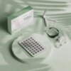 Na zdjęciu znajduje się biały panel LED z wieloma diodami, opakowanie z napisem "Nanoleaf Red Light Therapy Panel", składany stojak, czarna opaska oraz biały zasilacz z kablem. Wszystkie elementy są umieszczone na jasnym, zielonym tle.