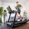 Na zdjęciu znajduje się bieżnia elektryczna w nowoczesnym wnętrzu z drewnianą podłogą, w tle widoczna jest szara sofa oraz regały z książkami i roślinami. Produkt to "Bieżnia elektryczna KETTLER Track 600 2206.5W 20 km/h".