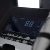 Zbliżenie na wyświetlacz orbitreka magnetycznego, na którym widoczne są cyfry i napisy: "TIME 5:36", "SPEED 8.8", "DIST. 0.1 KM", "CAL 2", oraz graficzne wykresy programów treningowych. Wyświetlacz jest podświetlony, a obok niego znajduje się fragment uch