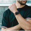 Mężczyzna w czarnej koszulce z kolorowymi paskami na rękawach nosi smartwatch z brązowym paskiem na nadgarstku. Na ekranie zegarka widoczne są ikony aplikacji i powiadomienia.