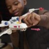 Na zdjęciu widoczna jest biała figurka statku kosmicznego z serii Star Wars z niebieskimi i żółtymi detalami oraz czerwonymi elementami. W tle znajduje się osoba trzymająca statek, a na pierwszym planie widoczny jest różowy pocisk.