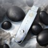 Na zdjęciu znajduje się urządzenie do peelingu kawitacyjnego z metaliczną powierzchnią, na której widoczne są krople wody oraz złoty wzór. W tle znajdują się gładkie, czarne kamienie, a na urządzeniu widoczny jest napis "LUMINEO".