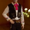 Lalka ubrana jest w elegancki strój z ciemną kamizelką ozdobioną złotymi wzorami, białą koszulę z kolorowymi haftami na kołnierzu i mankietach oraz różową wstążkę pod szyją. Na tle widoczne są rozmyte światła, przypominające świeczki.