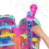 Kolorowy domek do zabawy z wieloma poziomami i akcesoriami, w tym figurkami, mebelkami i dekoracjami, z widoczną ręką dziecka manipulującą elementami. W tle unoszą się kolorowe konfetti, dodając wrażenie radości i zabawy.