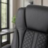 Czarny, ergonomiczny fotel biurowy z pikowanym wzorem na oparciu, widoczny na tle wnętrza z dużym oknem i rośliną w doniczce. Materiał fotela wygląda na skórzany lub imitujący skórę.