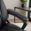 Czarny, ergonomiczny fotel biurowy obrotowy z widocznym podłokietnikiem i pikowanym oparciem, ustawiony w jasnym pomieszczeniu z biurkiem i rośliną w tle. Na biurku znajduje się zamknięty laptop.