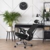 Na zdjęciu znajduje się czarne krzesło obrotowe o ergonomicznym kształcie, ustawione przed biurkiem z lampką i rośliną w doniczce. Na ścianie wisi okrągły zegar z białymi wskazówkami i cyframi na czarnym tle.