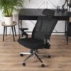 Czarne krzesło obrotowe o ergonomicznym designie stoi na drewnianej podłodze, obok czarnego biurka z rośliną w białej doniczce na małym stoliku. W tle widoczna jest biała ściana z dekoracyjnymi elementami.