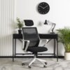 Na zdjęciu znajduje się czarno-białe krzesło obrotowe o ergonomicznym designie, ustawione przed czarnym biurkiem, na którym stoi lampa biurkowa i roślina w doniczce. Na ścianie wisi okrągły zegar z białymi cyframi na czarnym tle, a obok biurka znajdują si