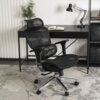 Czarne, ergonomiczne krzesło obrotowe znajduje się przed biurkiem z lampką i otwartym notesem. W tle widoczna jest roślina oraz regał z czarnymi pojemnikami.