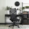 Czarne krzesło obrotowe o ergonomicznym kształcie stoi przed biurkiem, na którym znajduje się lampa biurkowa i roślina w wazonie. Na ścianie wisi okrągły zegar, a obok biurka znajduje się regał z książkami, roślinami i pudełkami do przechowywania.
