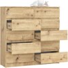 Drewniana komoda w kolorze dębu artisan z dziesięcioma szufladami, z których kilka jest wysuniętych. Na górze komody znajduje się biały talerz z białymi filiżankami i różowymi butami na obcasie.