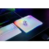 Na zdjęciu znajduje się biała podkładka z podświetleniem RGB, na której leży mysz komputerowa. Obok podkładki widoczna jest biała klawiatura z podświetleniem, a na podkładce znajduje się napis "RAZER".
