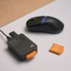Czarno-niebieska mysz bezprzewodowa z dwoma przyciskami bocznymi leży obok czarnego adaptera z pomarańczowym elementem i widocznymi napisami: "MOUSE", "BASE", "POLLING", "DPI" oraz logo producenta. Obok znajduje się pomarańczowa pokrywa z białymi oznaczen