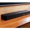 Czarny soundbar marki BOSE umieszczony na drewnianej powierzchni, z widocznym logo "BOSE" na przednim panelu. W tle znajduje się ściana pokryta jasnym, plecionym materiałem.