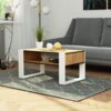 Stolik kawowy o wymiarach 92x53x45 cm z blatem w kolorze dąb craft i białymi matowymi frontami stoi na dywanie z geometrycznym wzorem. Na blacie znajdują się dwie dekoracyjne kule i doniczka z rośliną, a w tle widoczna jest szara sofa i lampa stojąca.