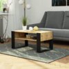 Stolik kawowy wykonany z drewna w kolorze dąb artisan z czarnymi matowymi frontami stoi na dywanie o geometrycznym wzorze, na którym znajdują się dekoracyjne przedmioty i roślina w doniczce. W tle widoczna jest szara sofa, lampa stojąca oraz fragment okna