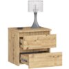 Na zdjęciu znajduje się szafka nocna w kolorze dąb artisan z dwoma wysuniętymi szufladami, na której stoi lampa z metalową podstawą i przezroczystym kloszem. Obok lampy leży mały, czarny przedmiot przypominający pilot.