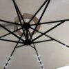 Beżowy parasol ogrodowy z oświetleniem LED, widziany od spodu, z czarną metalową konstrukcją wspierającą materiał. Światła LED są równomiernie rozmieszczone wzdłuż ramion konstrukcji.