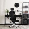 Na zdjęciu znajduje się czarno-białe krzesło obrotowe o ergonomicznym kształcie, ustawione przed biurkiem z lampką i rośliną w doniczce. W tle widać zegar ścienny oraz regał z książkami i segregatorami.