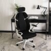 Czarno-białe krzesło obrotowe o ergonomicznym kształcie stoi na dywanie przed biurkiem, na którym znajduje się otwarta książka i lampa biurkowa. W tle widoczna jest roślina oraz pionowe żaluzje.