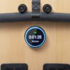Na zdjęciu widoczny jest wyświetlacz elektroniczny na drewnianej powierzchni, pokazujący czas "0:01:26" oraz prędkość "16.0 KM/H". Powyżej znajduje się uchwyt z czarną piankową osłoną i dwoma śrubami.