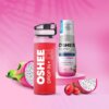 Na zdjęciu znajduje się butelka z czerwonym napojem z napisem "OSHEE DROP IN" oraz opakowanie koncentratu z etykietą "OSHEE DROP IN DI HYDROCONCENTRATE VITAMIN WATER ZERO VITAMINS + MINERALS". W tle widoczne są owoce smoczego owocu i czerwone winogrona na