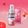 Na zdjęciu znajduje się butelka produktu "Hydrokoncentrat OSHEE DROP IN Vitamin Lemonade Malina i Grejpfrut 500 ml" obok szklanki z różowym napojem i kostkami lodu. W tle widoczne są maliny, plastry grejpfruta oraz rozpryskująca się woda, a na butelce wid