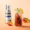 Na zdjęciu widoczna jest butelka produktu "OSHEE DROP IN" z napisem "HYDROCONCENTRATE VITAMIN TEA ZERO" oraz szklanka z napojem, kostkami lodu, plasterkiem brzoskwini i listkami mięty. Obok znajdują się kawałki brzoskwini i całe owoce na pomarańczowym tle