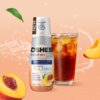 Butelka produktu "Hydrokoncentrat OSHEE DROP IN ZERO Vitamin Ice Tea Brzoskwinia 500 ml" stoi obok szklanki z napojem, na tle brzoskwiniowych elementów graficznych i rozprysków wody. Na etykiecie widnieje tekst: "NEW, koncentrat aż do 10L napoju*, PEACH F