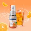 Butelka produktu "Hydrokoncentrat OSHEE DROP IN Vitamin Lemonade Pomarańcza 500 ml" stoi obok szklanki z pomarańczowym napojem i kostkami lodu, na tle pomarańczowym z plastrami pomarańczy. Na etykiecie widnieje tekst: "NEW, koncentrat aż do 10L napoju*, O