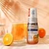 Butelka produktu "Hydrokoncentrat OSHEE DROP IN Vitamin Lemonade Pomarańcza 500 ml" stoi obok szklanki z napojem i plasterkami pomarańczy, na tle jasnej ściany z cieniami liści. Na etykiecie widnieje tekst: "NEW, koncentrat aż do 10L napoju, ORANGE FLAVOU
