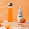 Na zdjęciu znajduje się butelka z hydrokoncentratem OSHEE DROP IN Vitamin Lemonade Pomarańcza 500 ml, obok której leży plasterek pomarańczy i kostki lodu. Tło jest pomarańczowe z delikatnym wzorem, a na butelce widnieje napis: "NEW koncentrat aż do 10L na